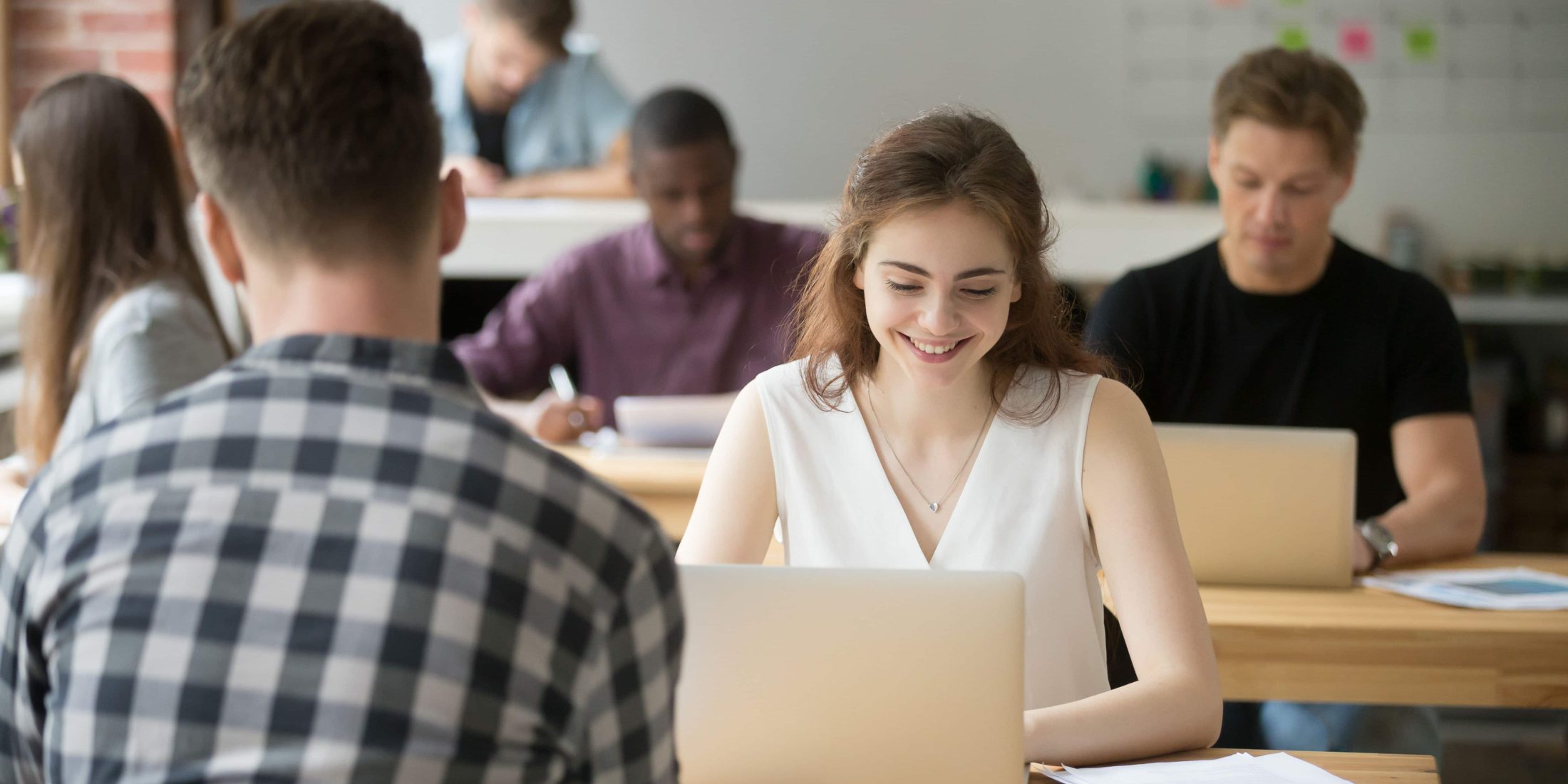 Young smiling woman working on laptop in coworking office, happy employee using computer sitting at desk in team shared space, attractive female student or client sales manager using pc at workplace